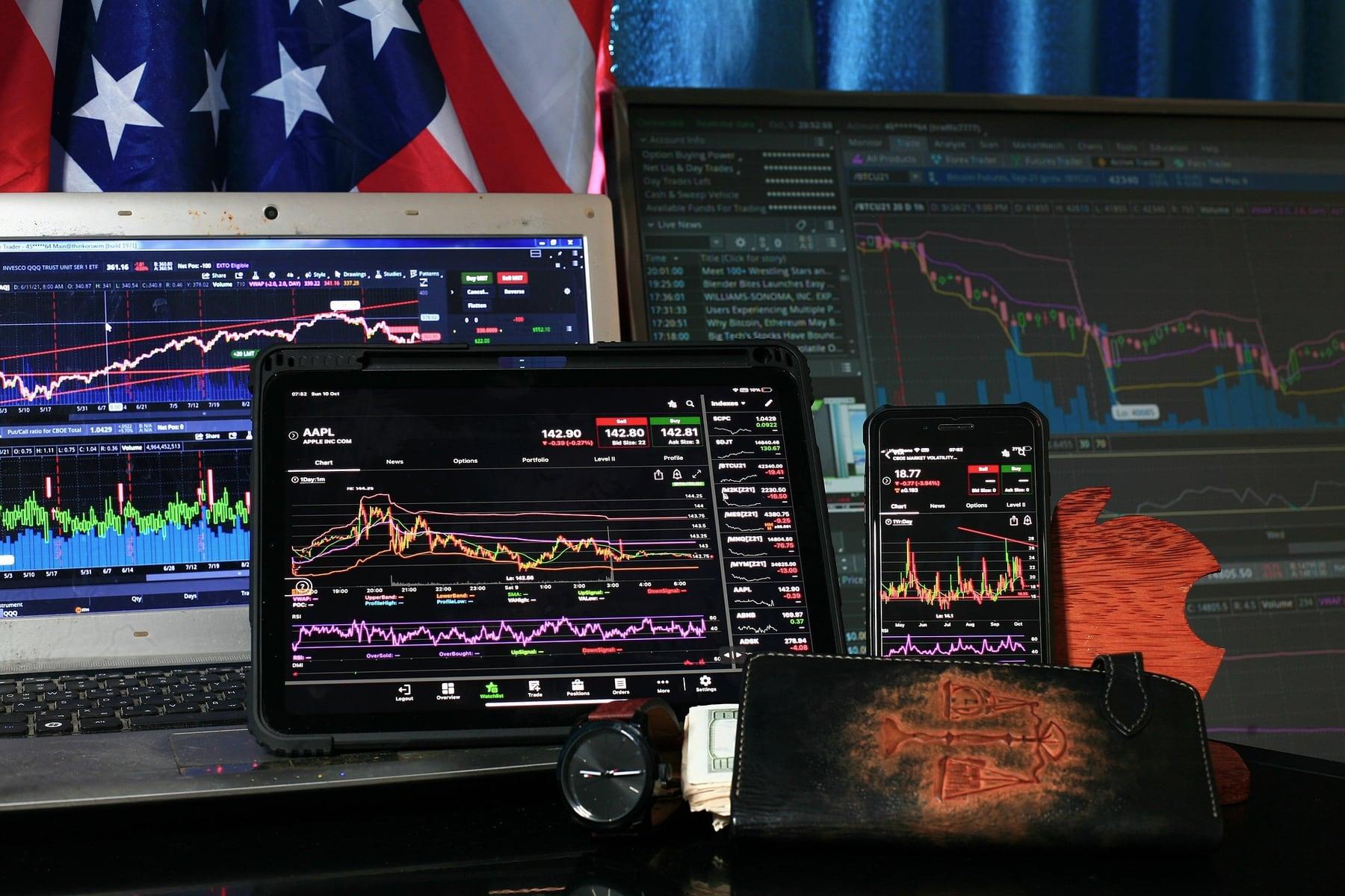 Trading desk with many monitors showing price charts and a US flag in the background