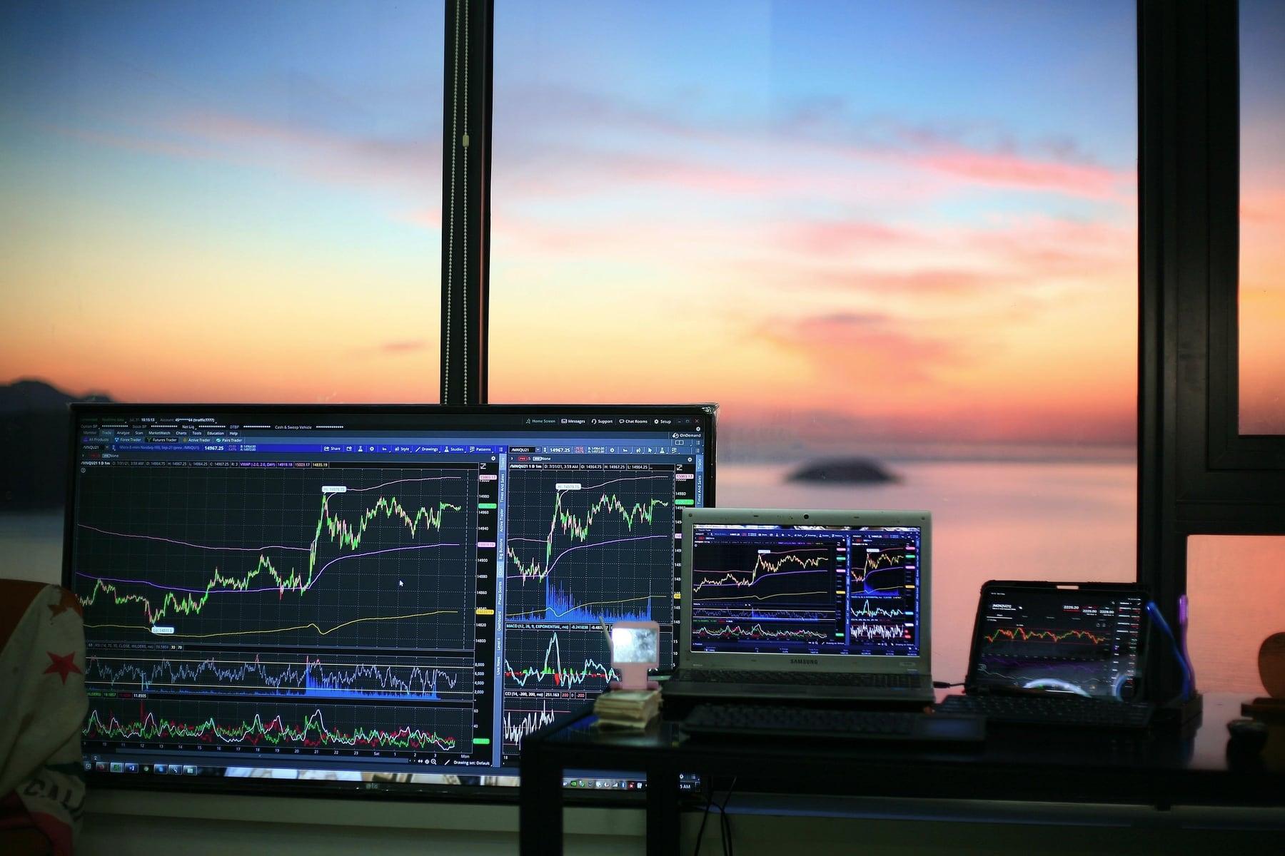 Computer monitors showing price charts in front of a window at sunset