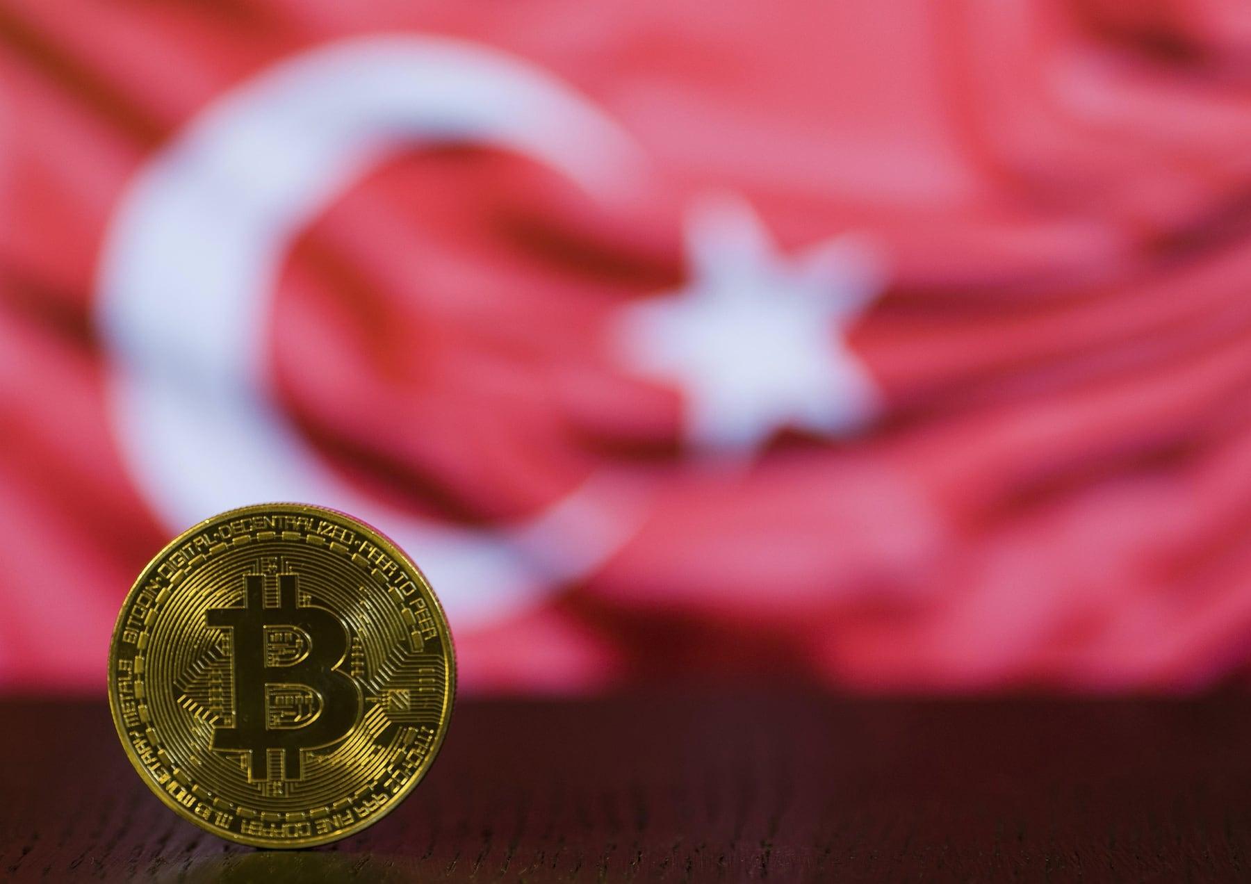 A gold bitcoin coin standing in front of a red Turkish flag with the white crescent and star